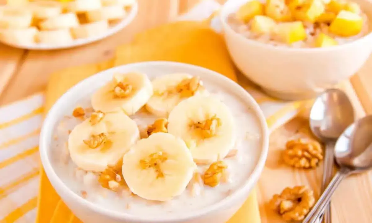 Un bol de talbina recette, garni de rondelles de banane et de noix. Une autre assiette de fruits est visible en arrière-plan.