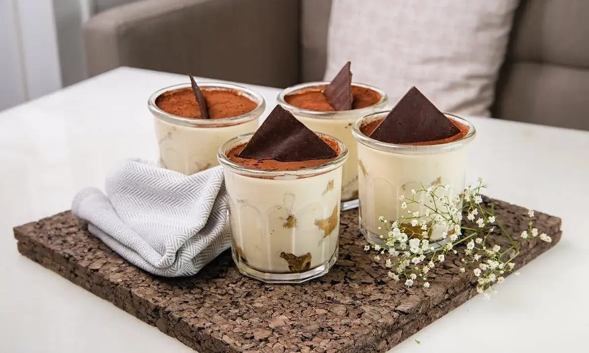 Quatre verrines de tiramisu, garnies de cacao et d'une feuille de chocolat. Une serviette grise et des petites fleurs blanches décorent le plateau en liège. Pourquoi mon tiramisu est liquide ?