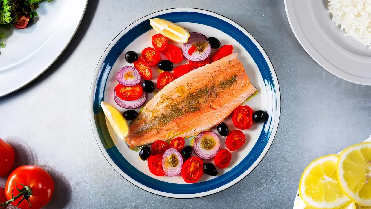 Une assiette colorée présente une truite au four, accompagnée de tomates cerises, d'oignons rouges, d'olives noires et de quartiers de citron.
