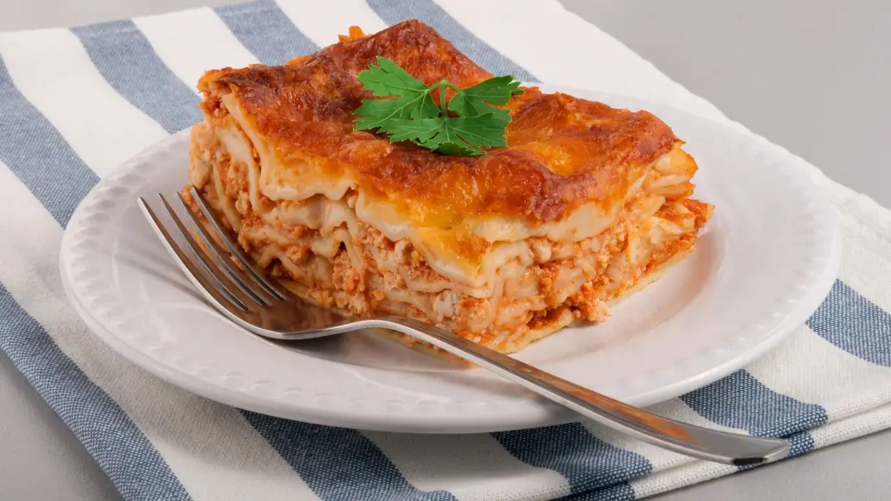 Une part de lasagne maison, dorée et appétissante, garnie d'une feuille de persil, sur une assiette blanche avec une fourchette.