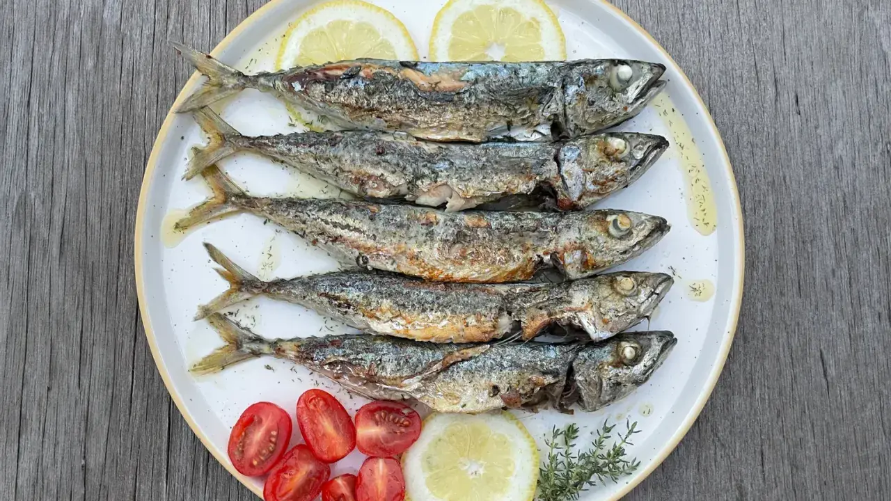 Cinq chinchards poissons grillés, servis sur une assiette avec des quartiers de citron, des tomates cerises et des herbes.