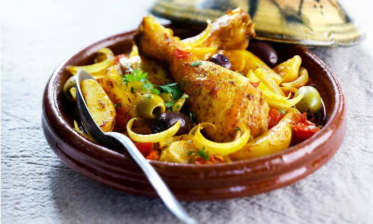 Un plat de tajine poulet citron, avec des morceaux de poulet dorés, des olives, des oignons et des tomates, servi dans un plat en terre cuite.