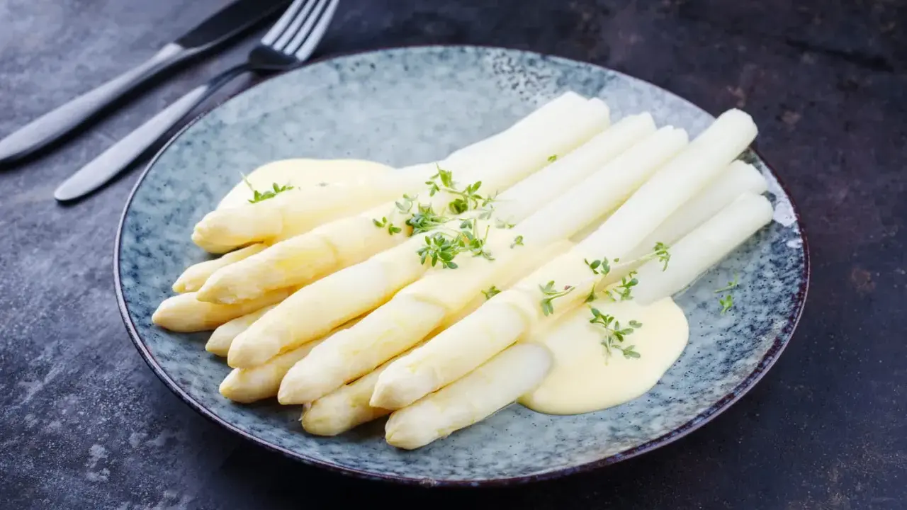 Asperges blanches nappées de sauce hollandaise et garnies d'herbes fraîches sur une assiette bleue.