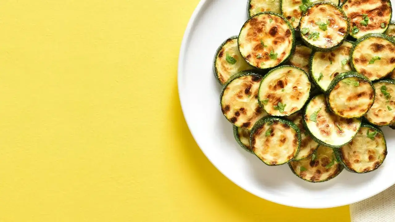 Courgettes rondes grillées à l'air fryer, parsemées d'herbes fraîches, sur une assiette blanche, sur fond jaune vif.