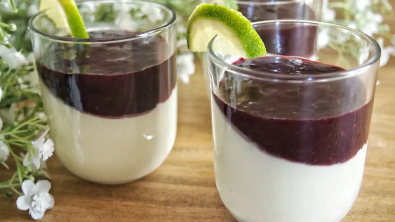 Trois panna cotta crémeuses, garnies d'un coulis pour panna cotta aux fruits rouges et d'une tranche de citron vert, sur un fond de fleurs blanches.