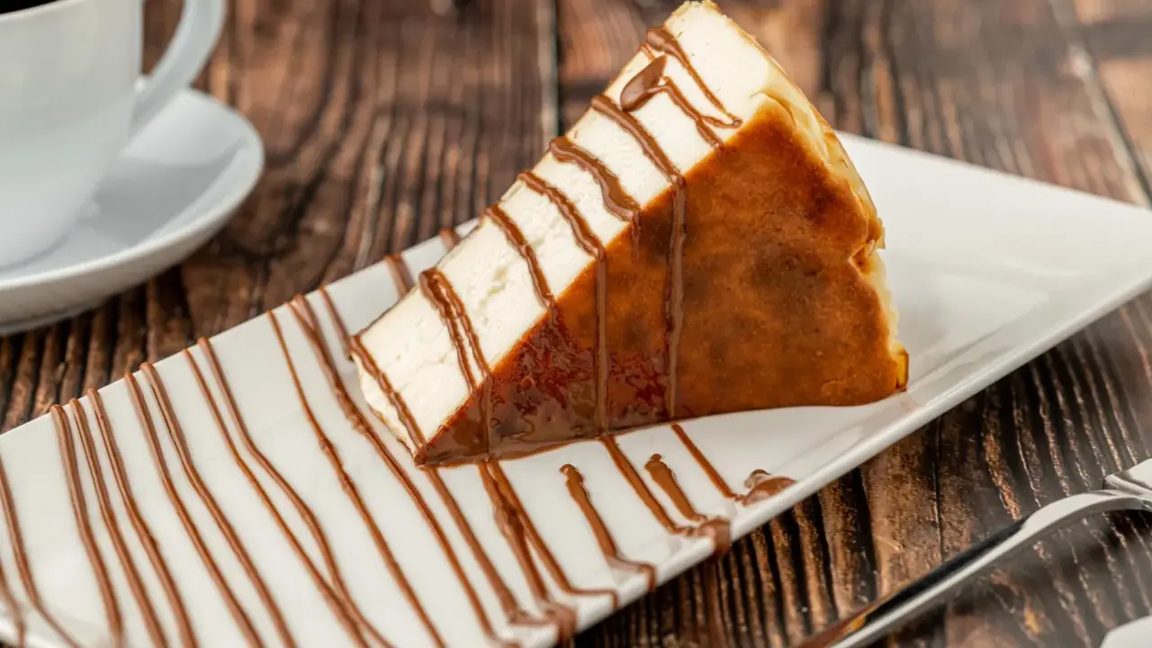 Tranche de san sebastian cheesecake, généreusement arrosée de sauce chocolat, sur une assiette blanche, avec une tasse de café en arrière-plan.