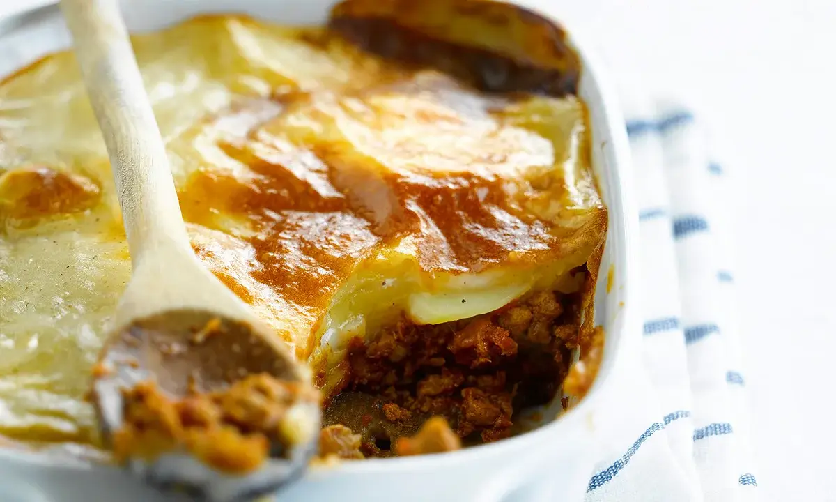 Une cuillère en bois sert une portion généreuse de moussaka végétarienne dorée et gratinée dans un plat blanc.