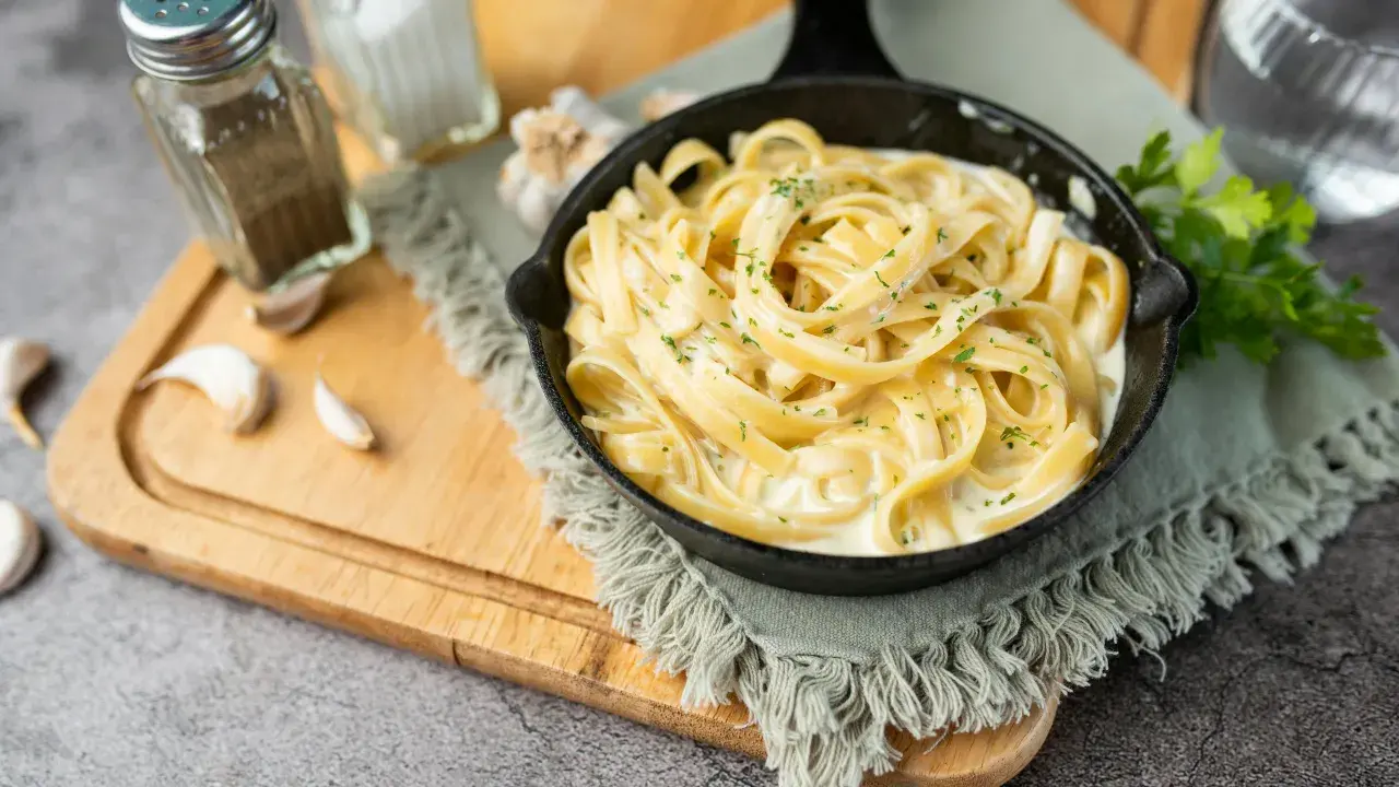 Un plat de fettuccine alfredo crémeux, garni de persil frais, servi dans une poêle en fonte. Des gousses d'ail et des moulins à sel et poivre sont disposés sur une planche à découper en bois.