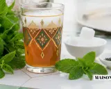 Un verre orné de motifs dorés et verts contient du thé marocain, accompagné de menthe fraîche, de sucre en morceaux et de feuilles de thé.