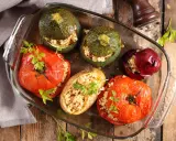Plat de farce à légume : tomates, courgettes rondes, oignon rouge et pomme de terre farcis de grains, joliment présentés dans un plat en verre.