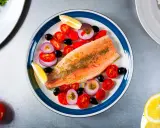 Une assiette colorée présente une truite au four, accompagnée de tomates cerises, d'oignons rouges, d'olives noires et de quartiers de citron.