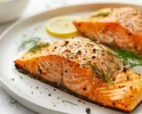 Deux pavés de saumon au four, assaisonnés de poivre et d'aneth, servis avec des tranches de citron sur une assiette blanche.