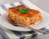 Une part de lasagne maison, dorée et appétissante, garnie d'une feuille de persil, sur une assiette blanche avec une fourchette.