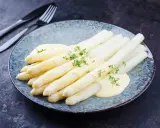 Asperges blanches nappées de sauce hollandaise et garnies d'herbes fraîches sur une assiette bleue.
