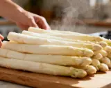 Une pile d'asperges blanches poêlées dégage de la vapeur sur une planche à découper en bois. Des mains ajustent délicatement les légumes.