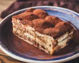 Tranche de gâteau tiramisu biscuit, généreusement saupoudrée de cacao, sur une assiette bleue.