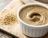 Un bol de tahini crémeux, prêt pour votre prochaine recette, à côté de graines de sésame sur une planche en bois.