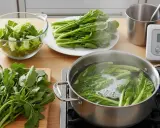 Préparation pour la cuisson d'épinards frais. Des légumes verts bouillonnent dans une casserole, d'autres attendent sur une assiette et une planche à découper.