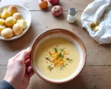 Bol de soupe crémeuse, garnie de fromage râpé et d'herbes. Une main tient le bol, à côté de pommes de terre et d'oignons. Le liquide de cuisson des pois chiches a servi de base.