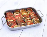 Un plat de tian de légumes, joliment arrangé en rondelles alternées de tomates, courgettes et aubergines, prêt à être dégusté.