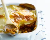 Une cuillère en bois sert une portion généreuse de moussaka végétarienne dorée et gratinée dans un plat blanc.
