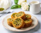Des rouleaux dorés de kadaif, c'est quoi ? Une pâtisserie croustillante aux pistaches, servie sur une assiette avec des roses en arrière-plan.