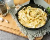 Un plat de fettuccine alfredo crémeux, garni de persil frais, servi dans une poêle en fonte. Des gousses d'ail et des moulins à sel et poivre sont disposés sur une planche à découper en bois.