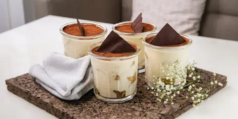 Quatre verrines de tiramisu, garnies de cacao et d'une feuille de chocolat. Une serviette grise et des petites fleurs blanches décorent le plateau en liège. Pourquoi mon tiramisu est liquide ?