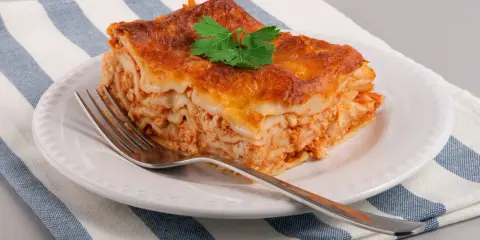 Une part de lasagne maison, dorée et appétissante, garnie d'une feuille de persil, sur une assiette blanche avec une fourchette.
