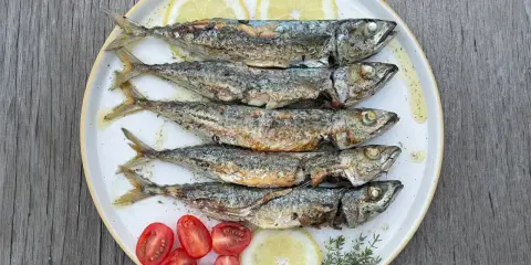 Cinq chinchards poissons grillés, servis sur une assiette avec des quartiers de citron, des tomates cerises et des herbes.