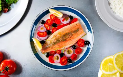 Une assiette colorée présente une truite au four, accompagnée de tomates cerises, d'oignons rouges, d'olives noires et de quartiers de citron.