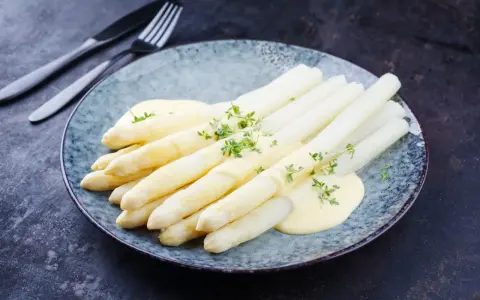 Asperges blanches nappées de sauce hollandaise et garnies d'herbes fraîches sur une assiette bleue.