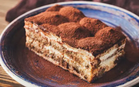 Tranche de gâteau tiramisu biscuit, généreusement saupoudrée de cacao, sur une assiette bleue.