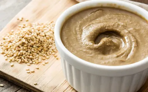 Un bol de tahini crémeux, prêt pour votre prochaine recette, à côté de graines de sésame sur une planche en bois.