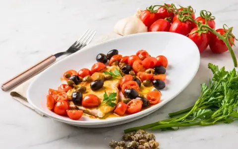 Filet de merlan servi avec tomates cerises, olives noires et câpres. Une assiette colorée et savoureuse.