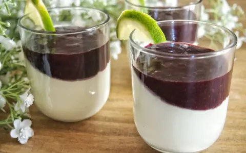 Trois panna cotta crémeuses, garnies d'un coulis pour panna cotta aux fruits rouges et d'une tranche de citron vert, sur un fond de fleurs blanches.