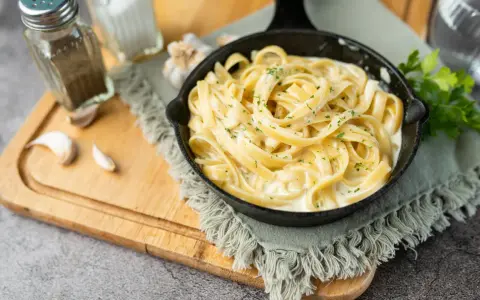 Un plat de fettuccine alfredo crémeux, garni de persil frais, servi dans une poêle en fonte. Des gousses d'ail et des moulins à sel et poivre sont disposés sur une planche à découper en bois.