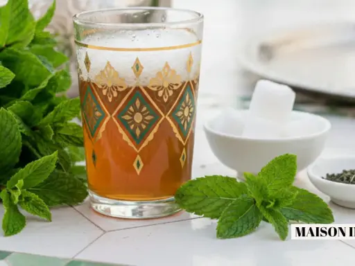 Un verre orné de motifs dorés et verts contient du thé marocain, accompagné de menthe fraîche, de sucre en morceaux et de feuilles de thé.