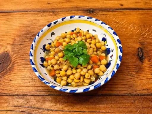 Un plat de pois chiches cuisinés avec des carottes et des oignons, garni de coriandre fraîche. La cuisson pois chiche est réussie, le plat est appétissant.