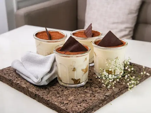 Quatre verrines de tiramisu, garnies de cacao et d'une feuille de chocolat. Une serviette grise et des petites fleurs blanches décorent le plateau en liège. Pourquoi mon tiramisu est liquide ?