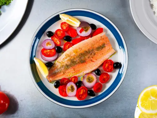 Une assiette colorée présente une truite au four, accompagnée de tomates cerises, d'oignons rouges, d'olives noires et de quartiers de citron.