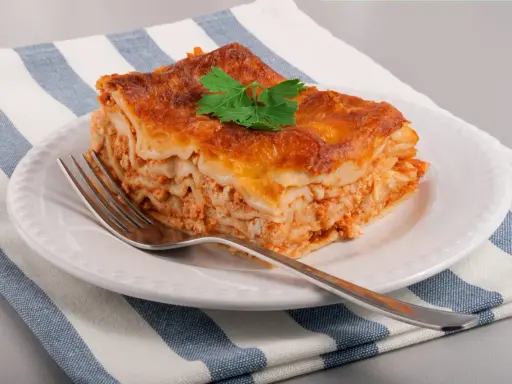 Une part de lasagne maison, dorée et appétissante, garnie d'une feuille de persil, sur une assiette blanche avec une fourchette.