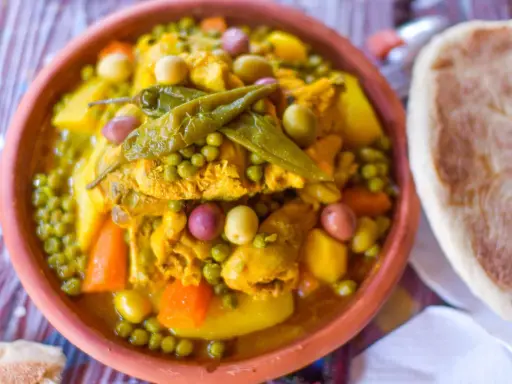 Un plat de tajine poulet aux olives, petits pois et carottes, garni de piments verts. Accompagné de pain.