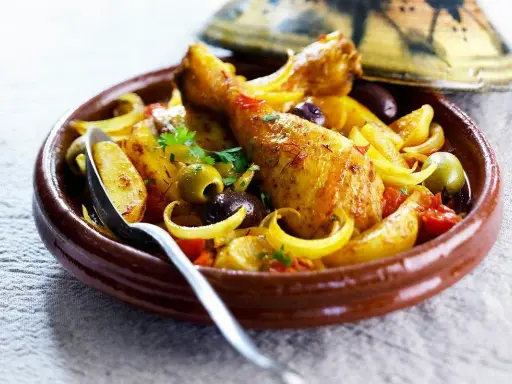 Un plat de tajine poulet citron, avec des morceaux de poulet dorés, des olives, des oignons et des tomates, servi dans un plat en terre cuite.