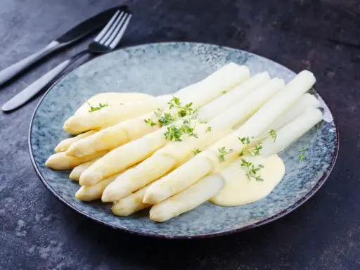 Asperges blanches nappées de sauce hollandaise et garnies d'herbes fraîches sur une assiette bleue.
