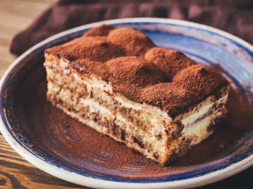 Tranche de gâteau tiramisu biscuit, généreusement saupoudrée de cacao, sur une assiette bleue.
