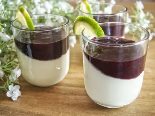 Trois panna cotta crémeuses, garnies d'un coulis pour panna cotta aux fruits rouges et d'une tranche de citron vert, sur un fond de fleurs blanches.