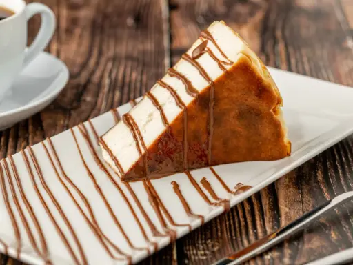 Tranche de san sebastian cheesecake, généreusement arrosée de sauce chocolat, sur une assiette blanche, avec une tasse de café en arrière-plan.