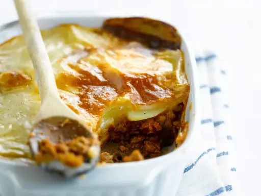Une cuillère en bois sert une portion généreuse de moussaka végétarienne dorée et gratinée dans un plat blanc.