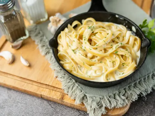 Un plat de fettuccine alfredo crémeux, garni de persil frais, servi dans une poêle en fonte. Des gousses d'ail et des moulins à sel et poivre sont disposés sur une planche à découper en bois.